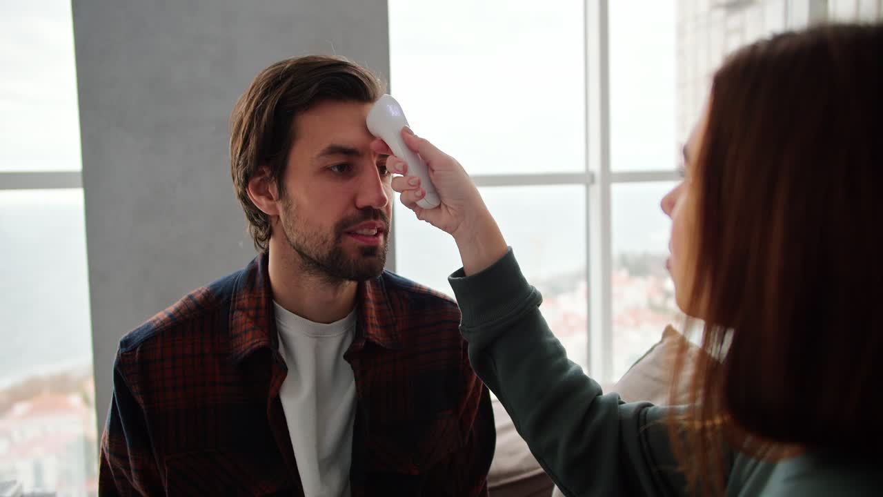 sobre su hombro una chica morena con un suéter verde mide la temperatura de su novio moreno usando un termómetro electrónico un tipo con paja en una camisa a cuadros habla de cómo tiene un resfriado mientras está sentado en el sofá en un apartamento moderno