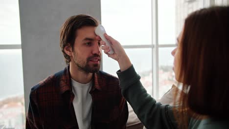 Por-Encima-Del-Hombro,-Una-Chica-Morena-Con-Un-Suéter-Verde-Mide-La-Temperatura-De-Su-Novio-Moreno-Usando-Un-Termómetro-Electrónico.-Un-Chico-Con-Barba-Incipiente-Y-Camisa-A-Cuadros-Habla-De-Cómo-Está-Resfriado-Mientras-Está-Sentado-En-El-Sofá-De-Un-Apartamento-Moderno.