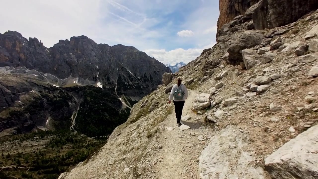 Giovane donna con uno zaino che fa escursioni su un sentiero stretto ed esposto nelle Alpi italiane. Vista panoramica delle cime rocciose e della profonda valle sottostante.