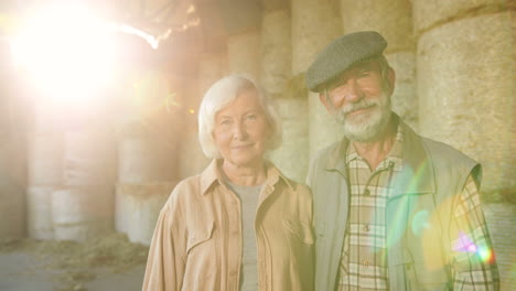 portrait of happy old caucasian married couple of farmers looking and smiling at camera while standing in stable with hay stocks