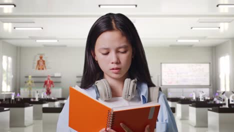 불만족한 아시아 십대 소녀 학생이 배을 들고 책을 읽고 과학 실험실에 서서 머리를 흔들고 있습니다.