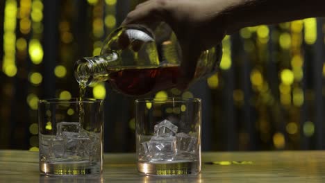 pouring of whiskey, cognac or brandy from bottle into glasses with ice cubes. shiny background