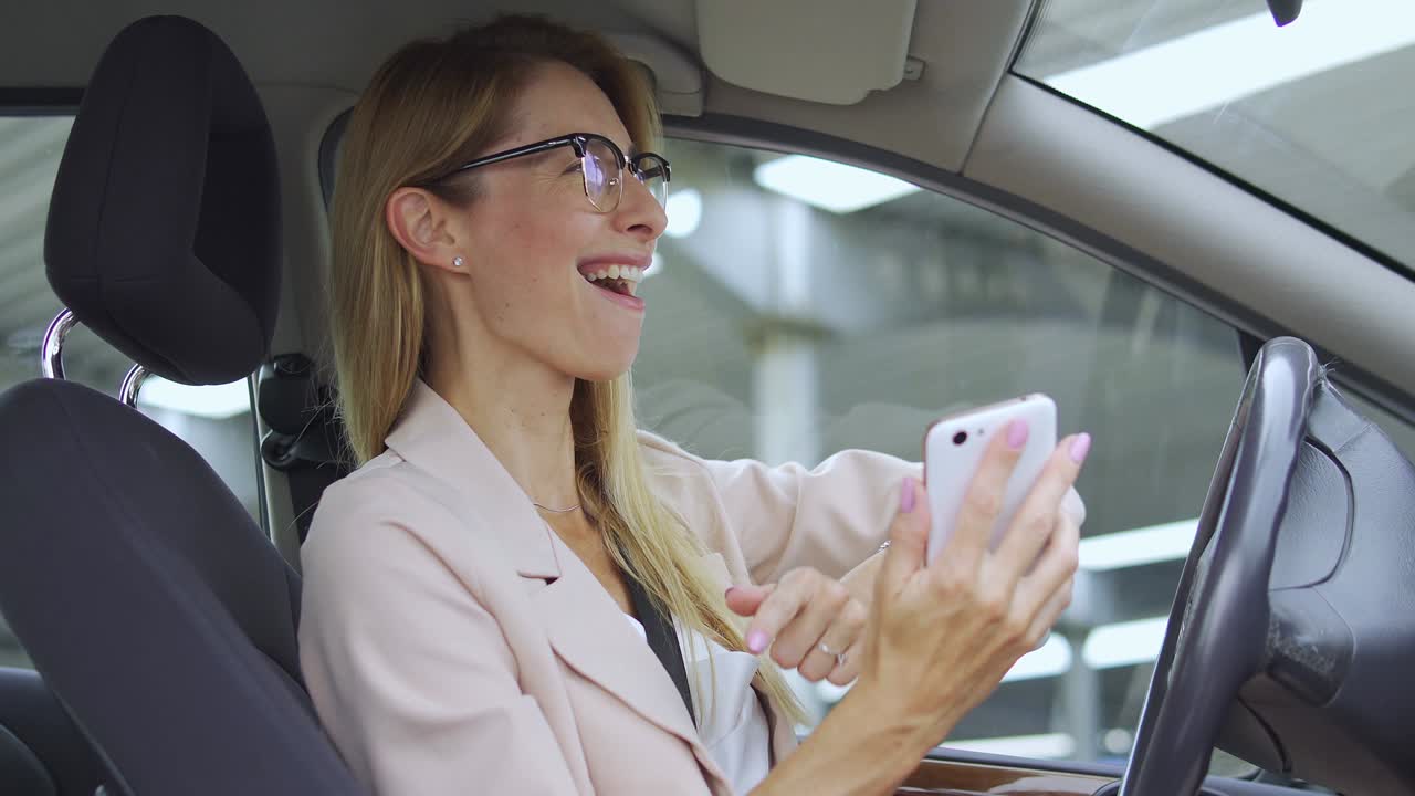 Beautiful excited blond woman in elegant light suit actively moving to rhythm of favourite song, listening to music on smartphone sitting in car, positive successful lady enjoying good news