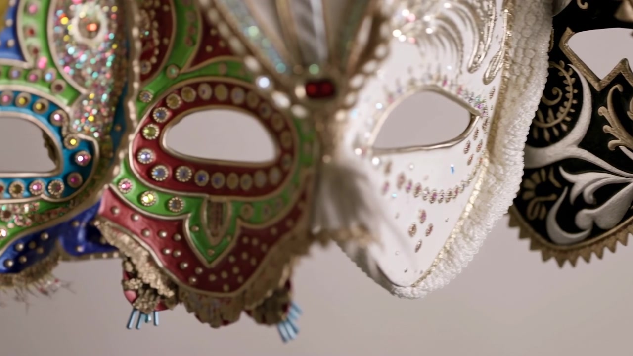 Close-up em camadas de máscaras de mascarada penduradas em azul, verde, vermelho, branco, preto e dourado, com acabamento em pedrarias, detalhes metálicos, bordas com penas e fundo suave desfocado para uma peça artesanal festiva e refinada.