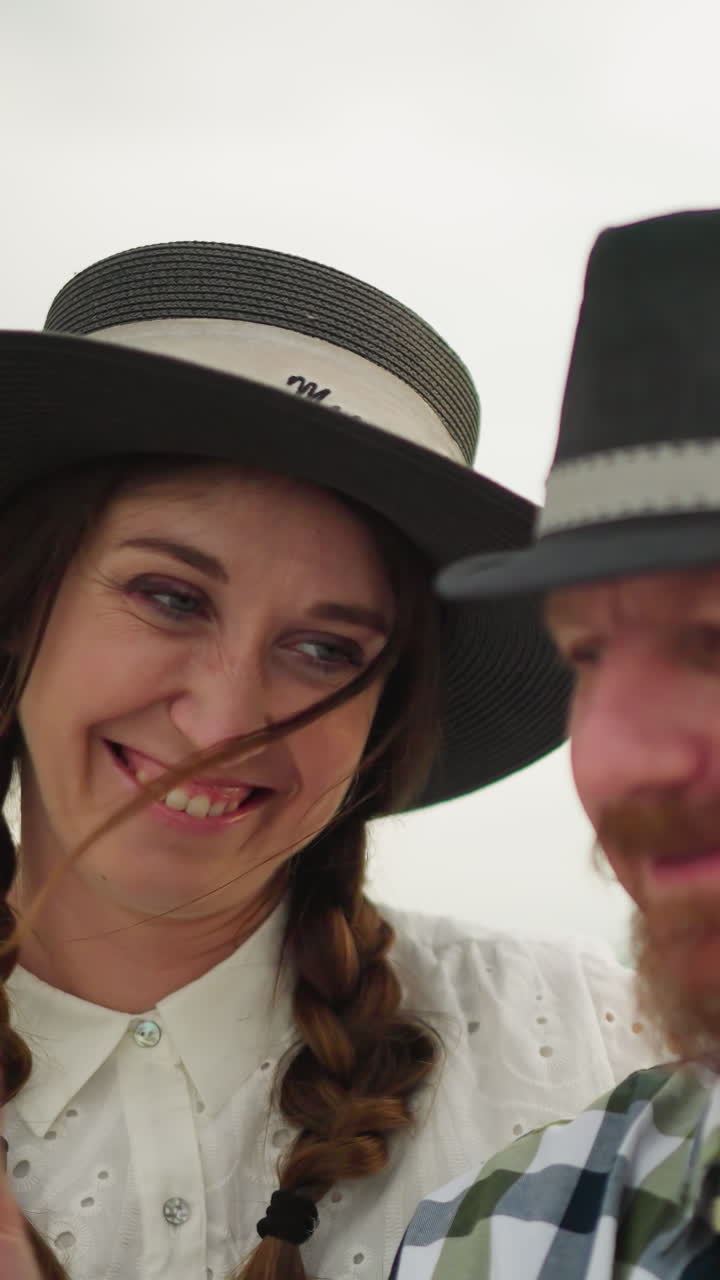 un primer plano de una pareja amorosa de pie juntos, ambos con sombreros. el hombre, en una camisa a cuadros, es tocado suavemente en la cara por un pedazo de hierba sostenido por la mujer en un vestido blanco