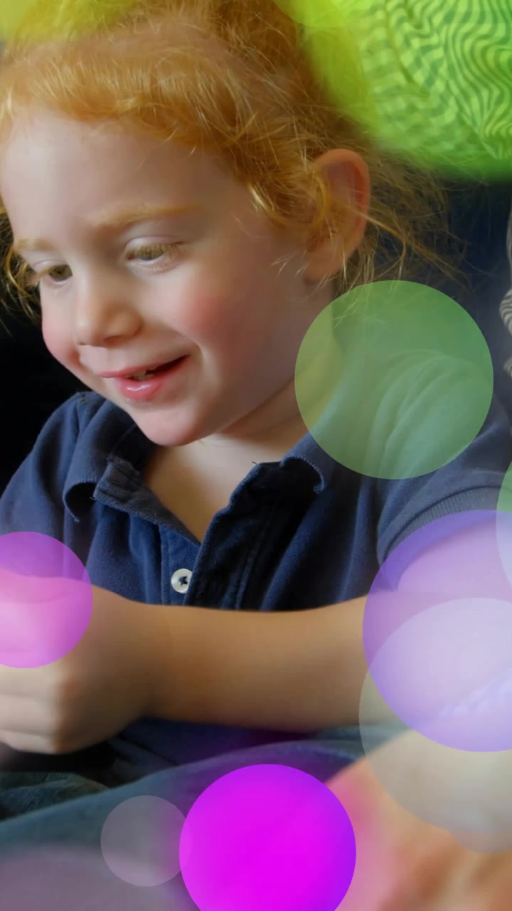 Video verticale: Bambino che tocca il telefono sul divano, punti colorati che fluttuano con un adulto che lo guida per il gioco tecnologico.