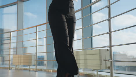 girl walking through sunlit modern hallway balancing novel on head with hand lifted for balance, wearing glasses, white shirt and black pants, framed by tall windows revealing urban view beyond
