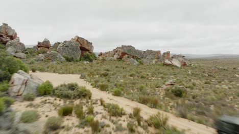 drone flies fast over desert landscape and through rock formations in cederberg wilderness area in south africa and flies over a road where a black off-road vehicle is driving