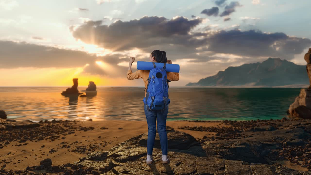 Vista traseira de corpo inteiro de uma caminhante com mochila de alpinismo gritando gol, celebrando o sucesso enquanto está de pé na praia durante o pôr do sol