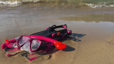 red and black fins mask and snorkel left on sandy beach with waves breaking on shore