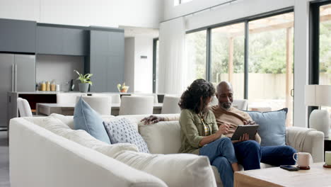 happy biracial couple sitting on couch and using tablet at home, slow motion