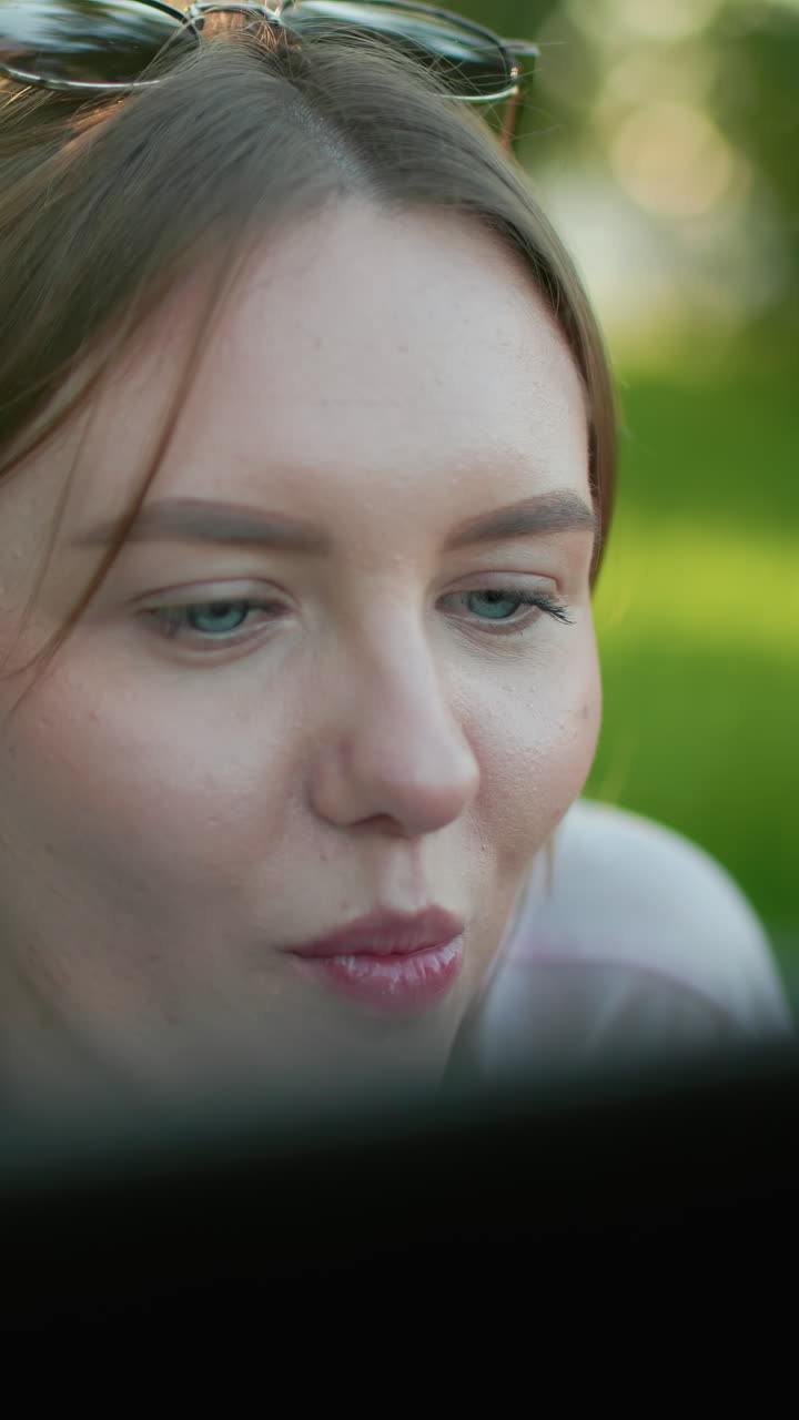White lady with glasses resting on head lying on grass smiling warmly while taking a bite from apple in her hand, enjoying relaxed outdoor moment, surrounded by greenery