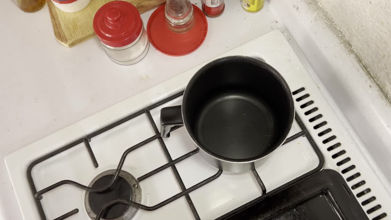 Close-up of hand pouring water from orange container into a pot on a gas stove in kitchen setting.
