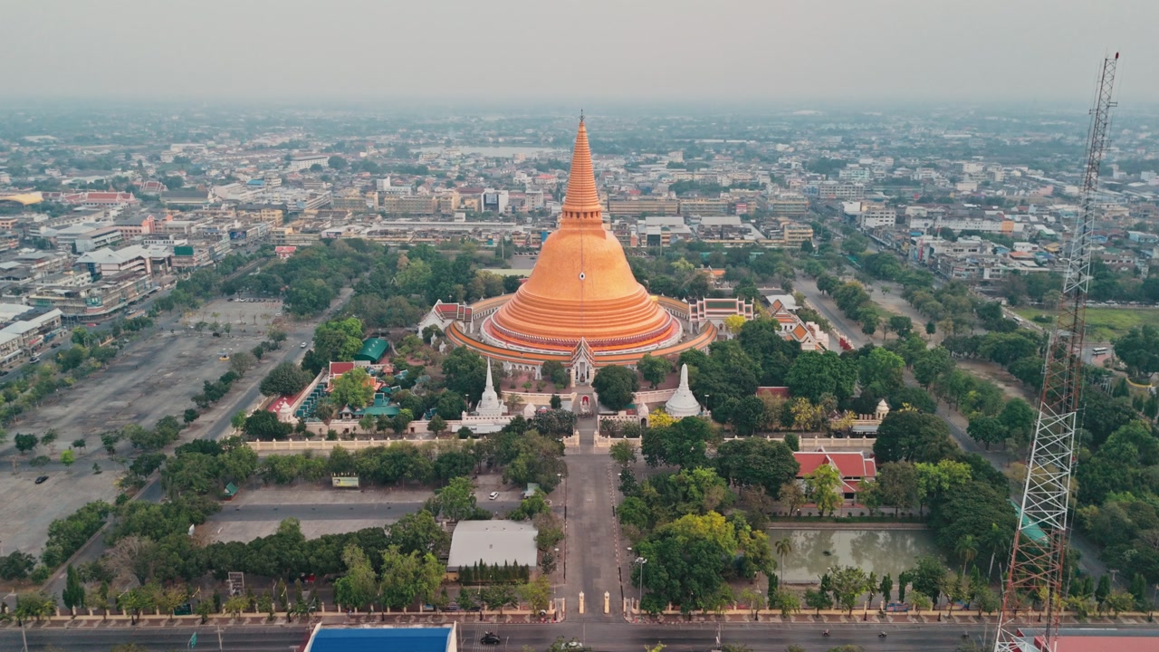 High angle drone perspective moves forward toward huge golden stupa rising above urban center while majestic Buddhist monument dominates hazy gray city skyline in Thailand during serene beautiful morn