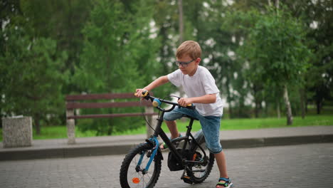 a young boy in a white t-shirt and denim shorts moves his bicycle and rode off, surrounded by greenery, benches, and a calm park environment