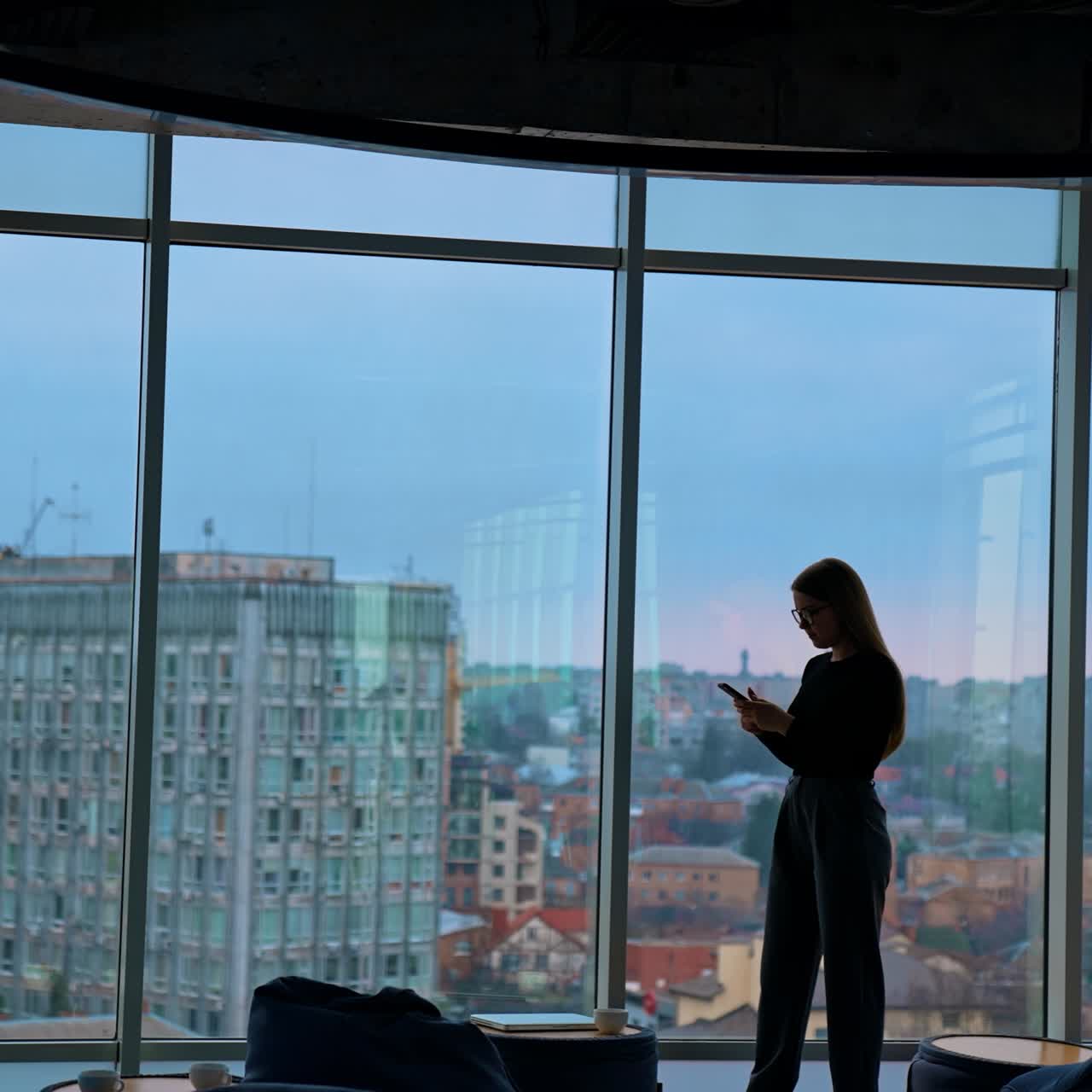 Full-length portrait on a business woman in modern office. Panoramic view on large office windows with cityscape indoors. Beautiful female business worker with a phone