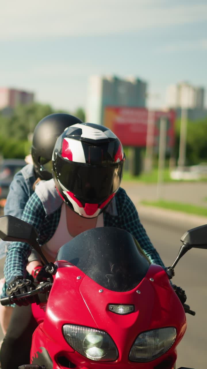 두 자매가 빨간색 파워 자전거를 타고, 둘 다 헬을 착용하고, 도시 도로를 항해하는 동안, 그녀의 천 뒤에 있는 여인은 바람에 날아다니며, 건물, 자동차, 그리고 초록색이 배경에 나타납니다.