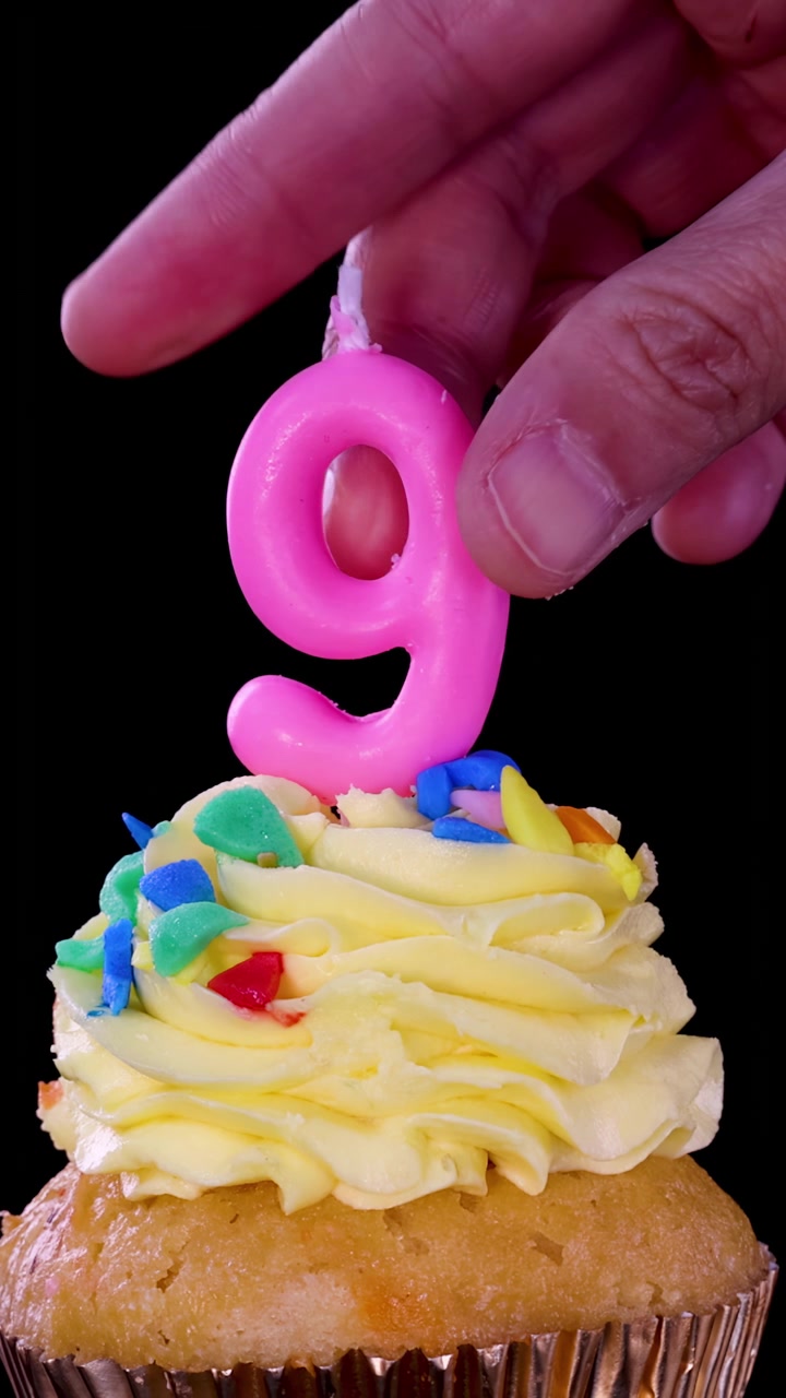 Una mano inserisce una candela rosa a forma di numero nove in un cupcake glassato colorato su sfondo nero, primo piano