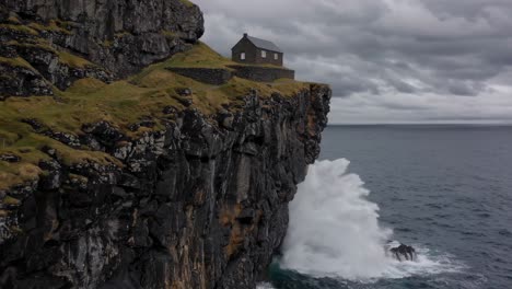 kallur lighthouse is standing on a cliff overlooking powerful ocean waves crashing against the rocks below, with a dramatic cloudy sky above in the faroe islands