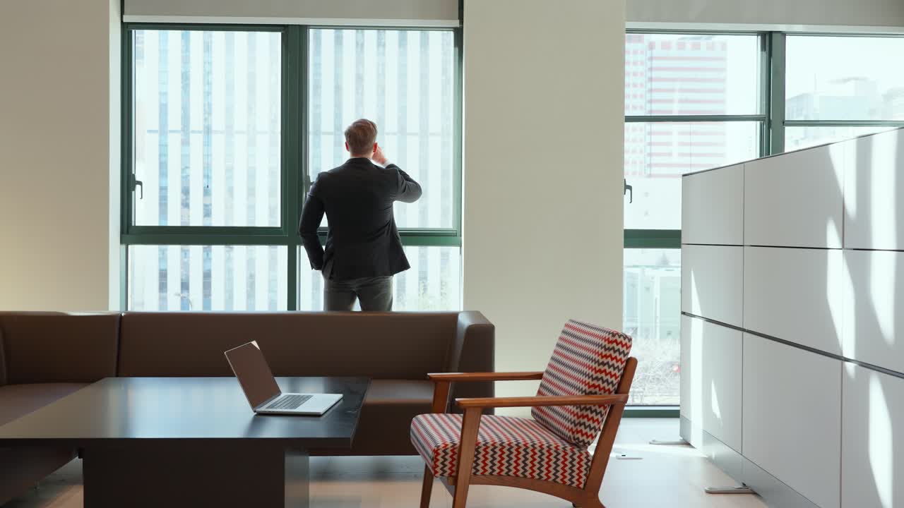 A person in a business suit stands contemplatively by large office windows drinking coffee in modern workspace with a wooden table and laptop, a retro-styled striped armchair, and white storage units