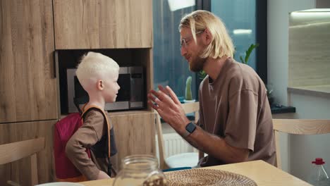 happy little albino boy with white hair color with a red backpack on his back communicates with his dad, a blond man with a beard and glasses in the kitchen after breakfast before the start of the school day at school