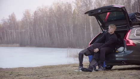 Ice-diver-sitting-in-car-boot-puts-on-drysuit-next-to-iced-lake