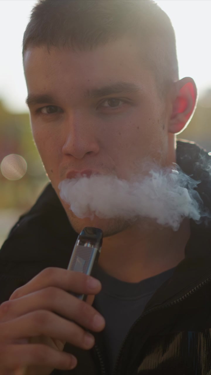 Close up of man holding and using electronic vaping device with visible vapor. subject wears casual clothing including jacket over t shirt in soft natural light that creates lens flare