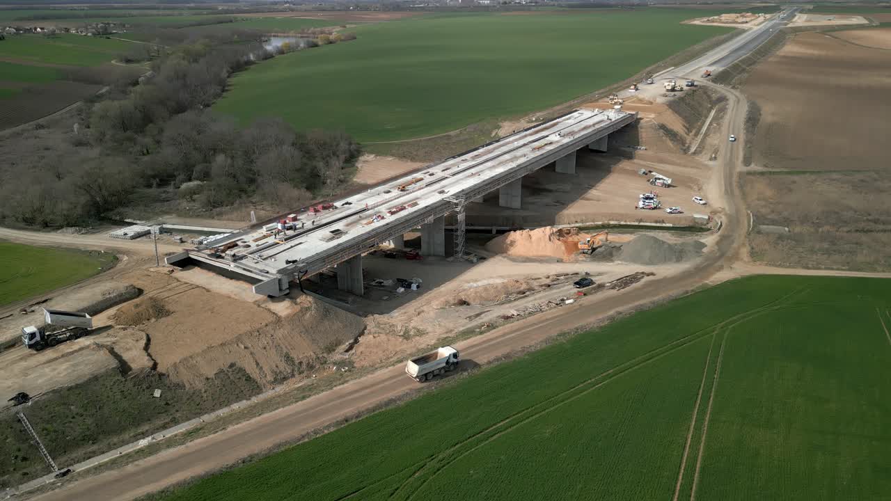 Aerial view of a bridge under construction in a rural landscape with vehicles and dirt paths visible