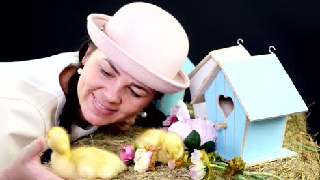 portrait, a pretty young woman with two pigtails and in a funny pink hat playing with small yellow ducklings. in the background a haystack, colored birdhouses, and flowers