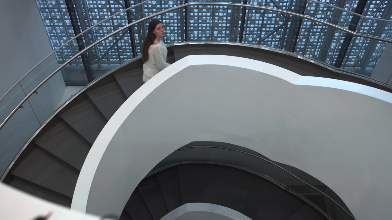Top-down view of modern interior with spiral staircase, curved white balustrade, dark recessed steps, and woman descending along upper curve beneath blue patterned glazing and shadow