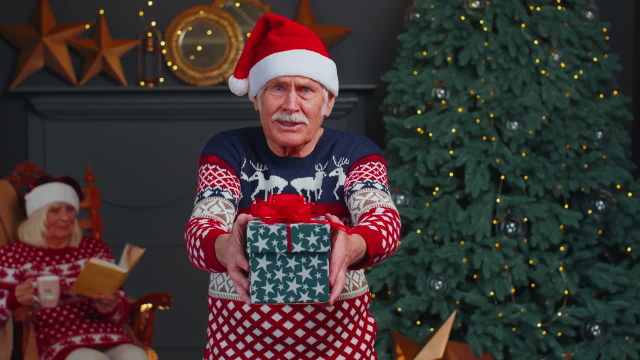 Mature grandfather in new year sweater and hat presenting christmas gift box holidays at home
