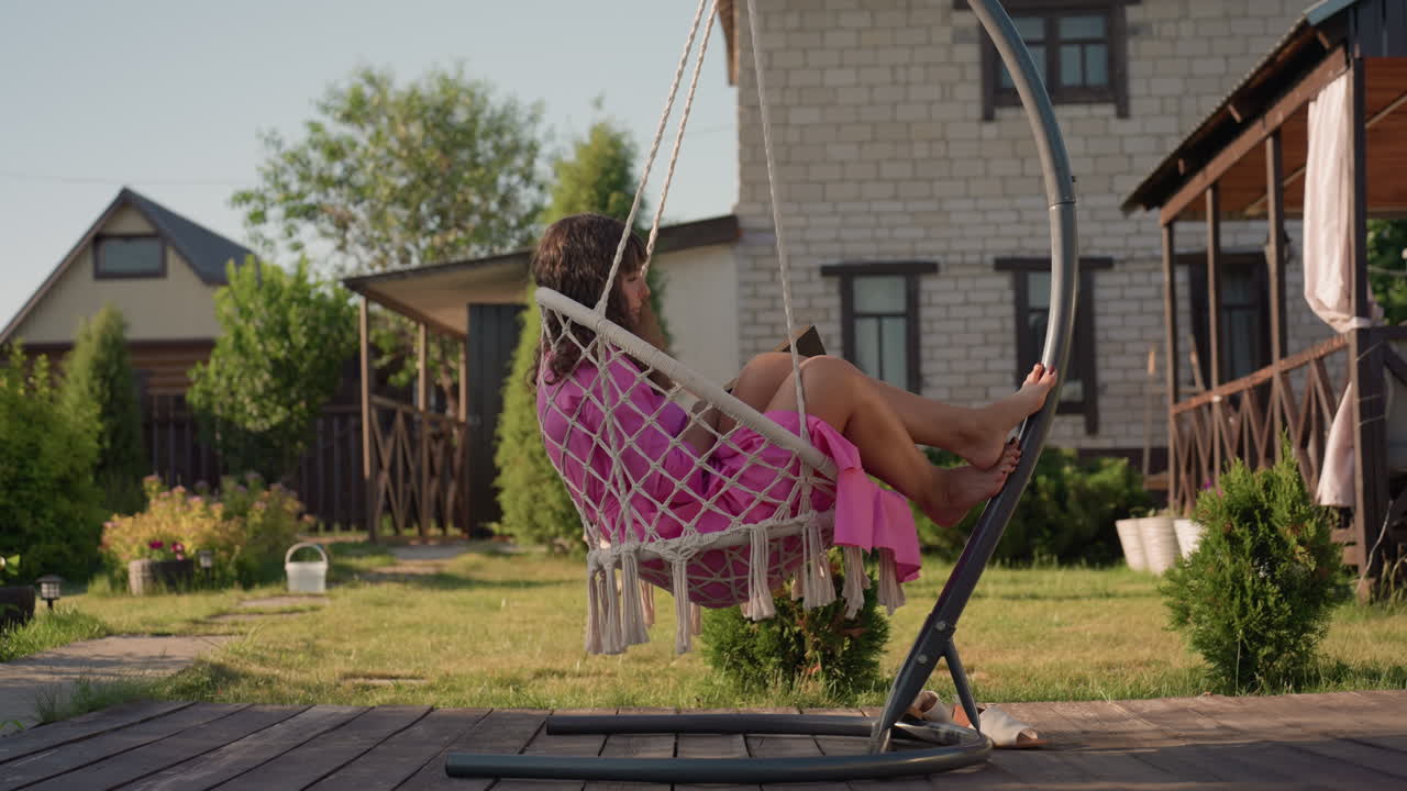 Leisurely Lady Resting In Sunny Backyard, Peaceful Woman Reclining In Woven Seat On Garden Patio, Serene Woman Unwinding On Wooden Deck Amid Blooming Cottage Garden In Warm Sunshine