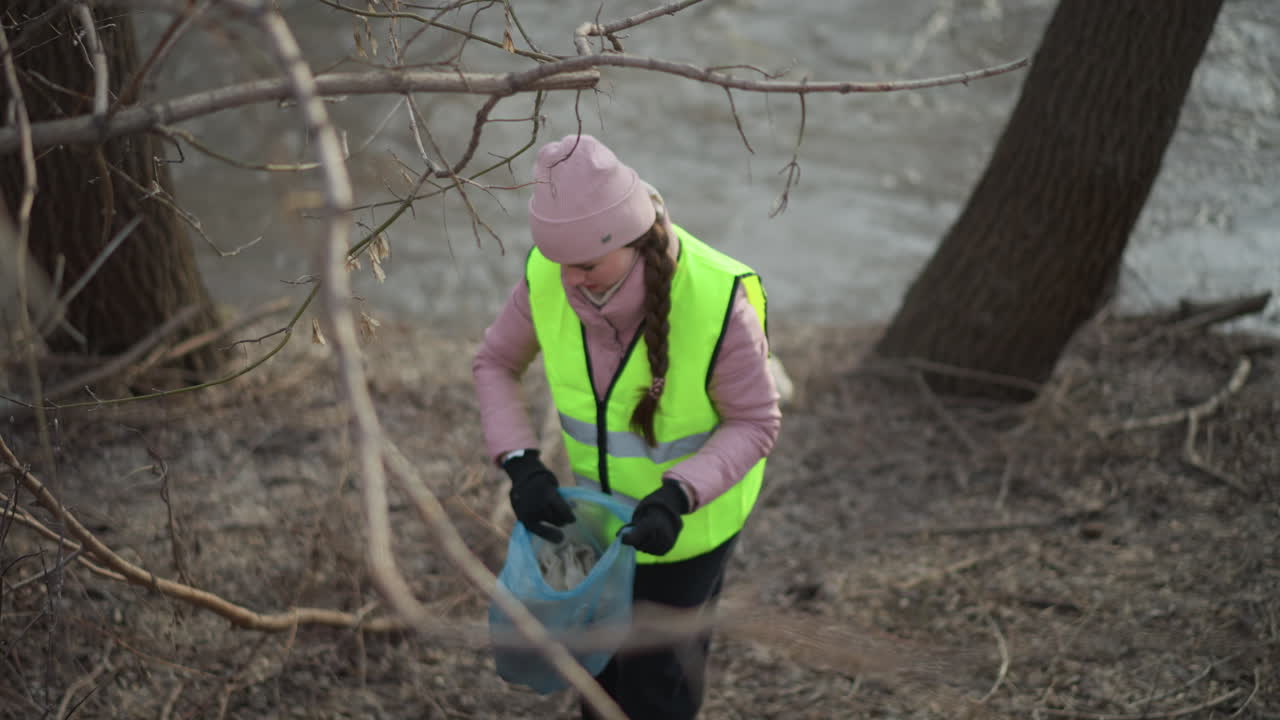 Woman in reflective vest and pink beanie bends down to collect trash in blue plastic bag among dry branches and debris on riverbank during environmental cleanup, removing waste to protect nature