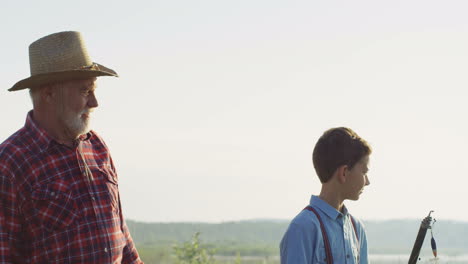 Caucasian-grandfather-and-his-little-grandson-walking-together-near-a-lake-while-holding-fishing-rods-in-the-morning