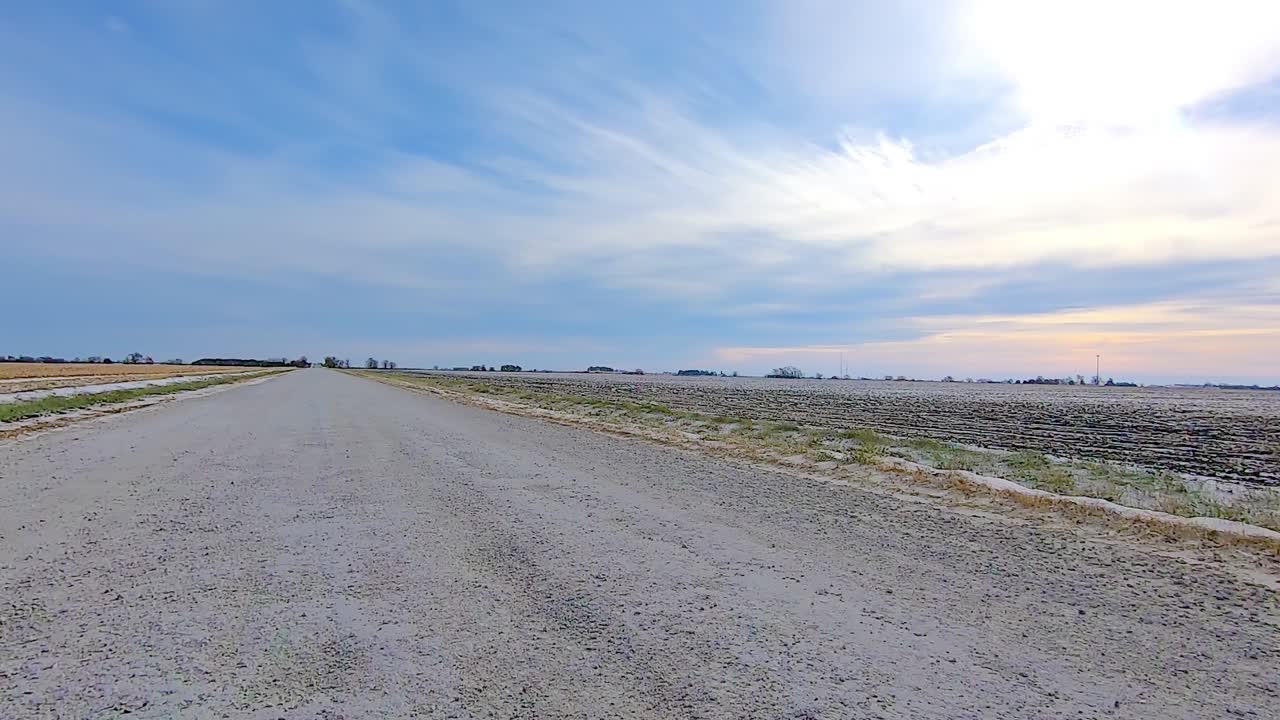 POV through the rear window while driving on a rural gravel road past empty fields on a sunny winter afternoon; there is a light dusting of snow in the ditches