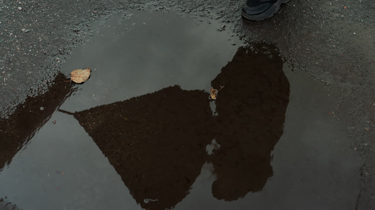 Reflektion i en vandpyt af en fodgænger med paraply, nærbillede ovenfra af våd asfalt og flydende blad, paraplysilhuet og sko, der går forbi, subtile krusninger og stemningsfuldt overskyet lys fremkalder