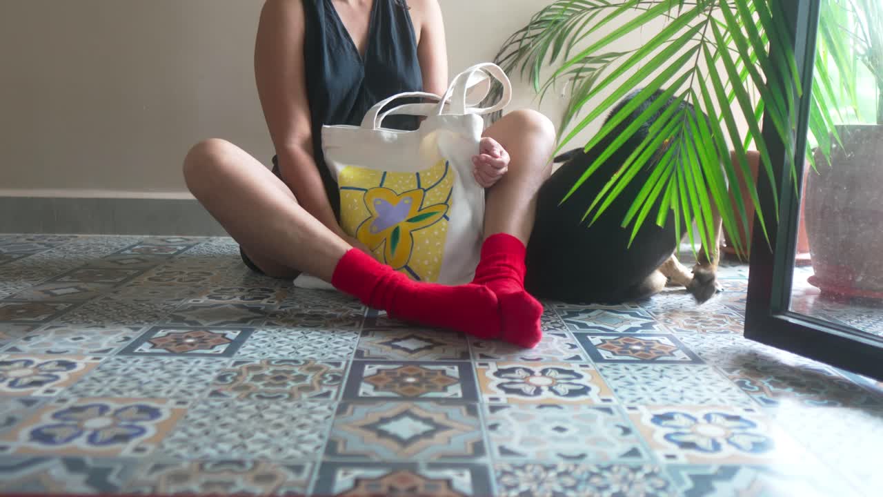Giovane donna seduta su un pavimento a fantasia in interni, tiene una borsa tote riutilizzabile con un design artistico colorato. La luce naturale dalla finestra e una pianta da interno creano una scena di vita moderna e tranquilla.