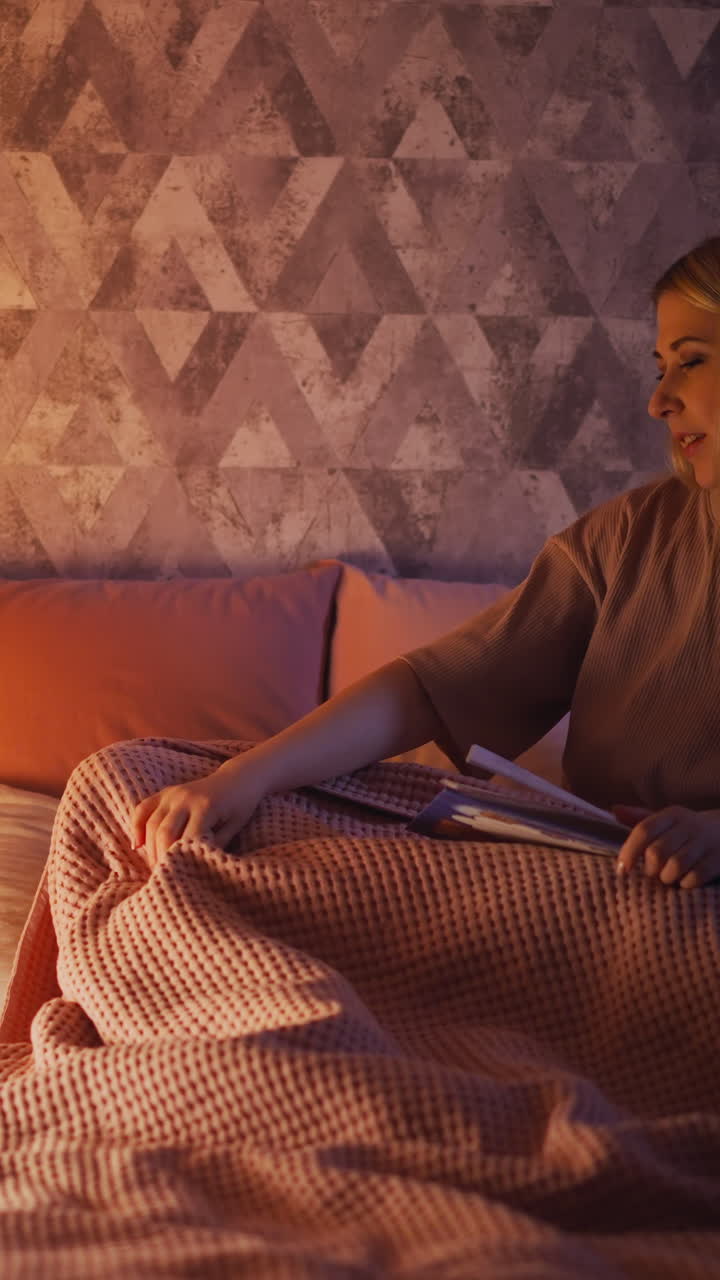Careful mommy with book adjusts pink blanket while child rolls over falling asleep near toy wigwam with glowing lamp in shady bedroom slow motion