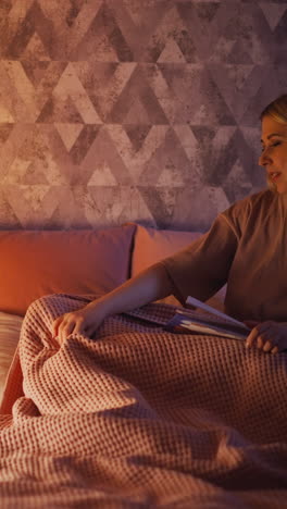 careful mommy with book adjusts pink blanket while child rolls over falling asleep near toy wigwam with glowing lamp in shady bedroom slow motion