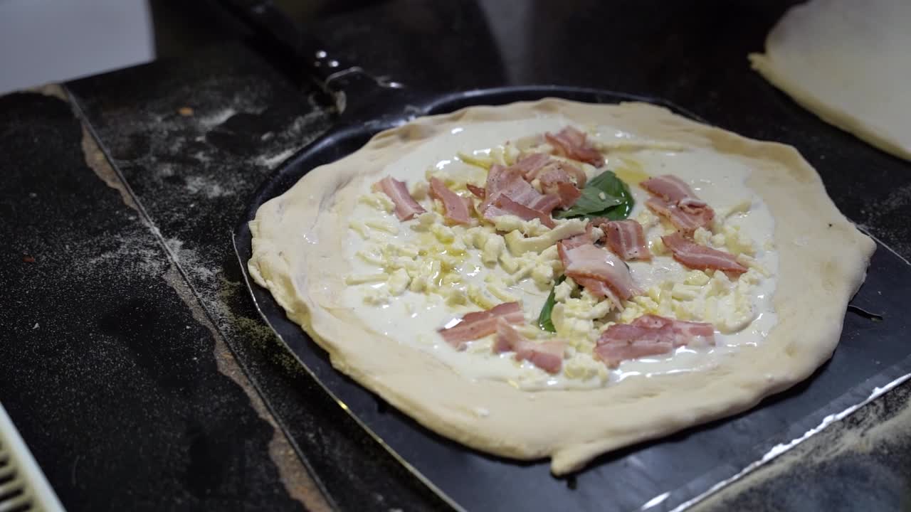 A chef's hands shaping pizza dough on a black baking tray, topped with a creamy white sauce, grated cheese, fresh basil, and slices of bacon. The process highlights the steps of making a gourmet pizza
