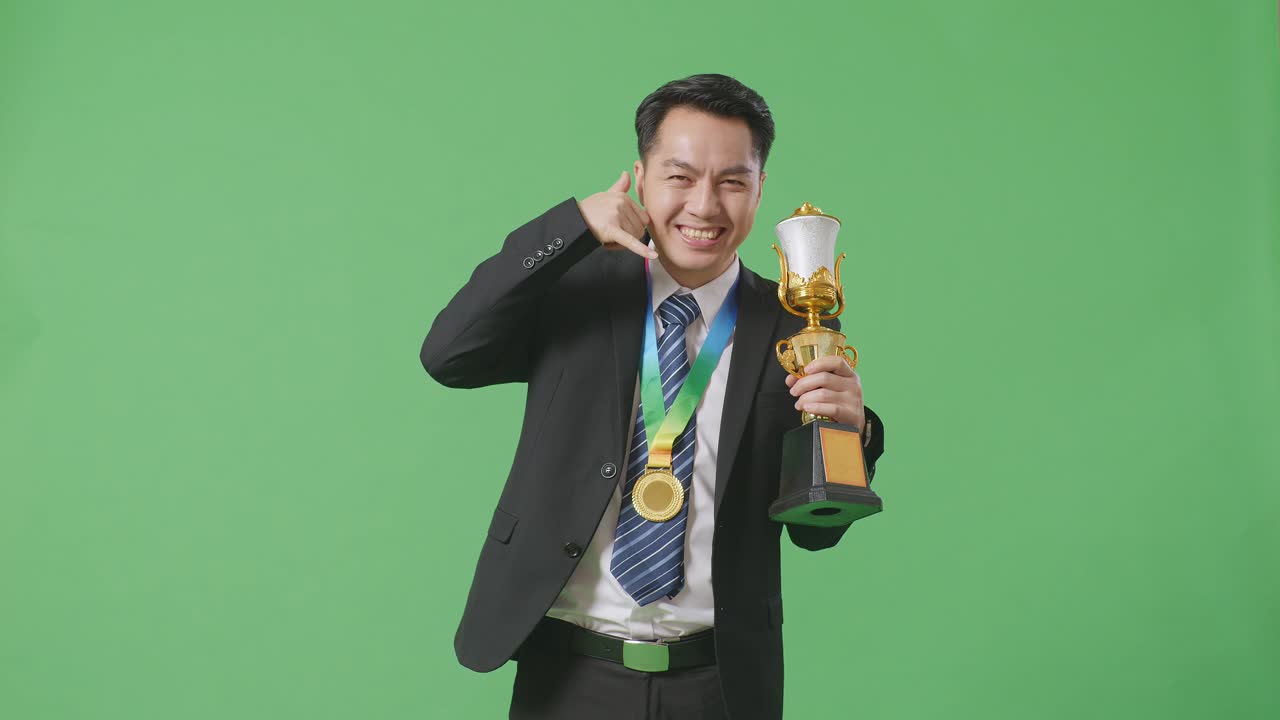 슈트와 타이를 입은 아시아 사업가, 금메달과 트로피를 들고, '나에게 전화해' 제스처를 하고, 카메라에 미소를 지으며, 스튜디오의 녹색 스크린 배경에서 첫 번째 우승자로