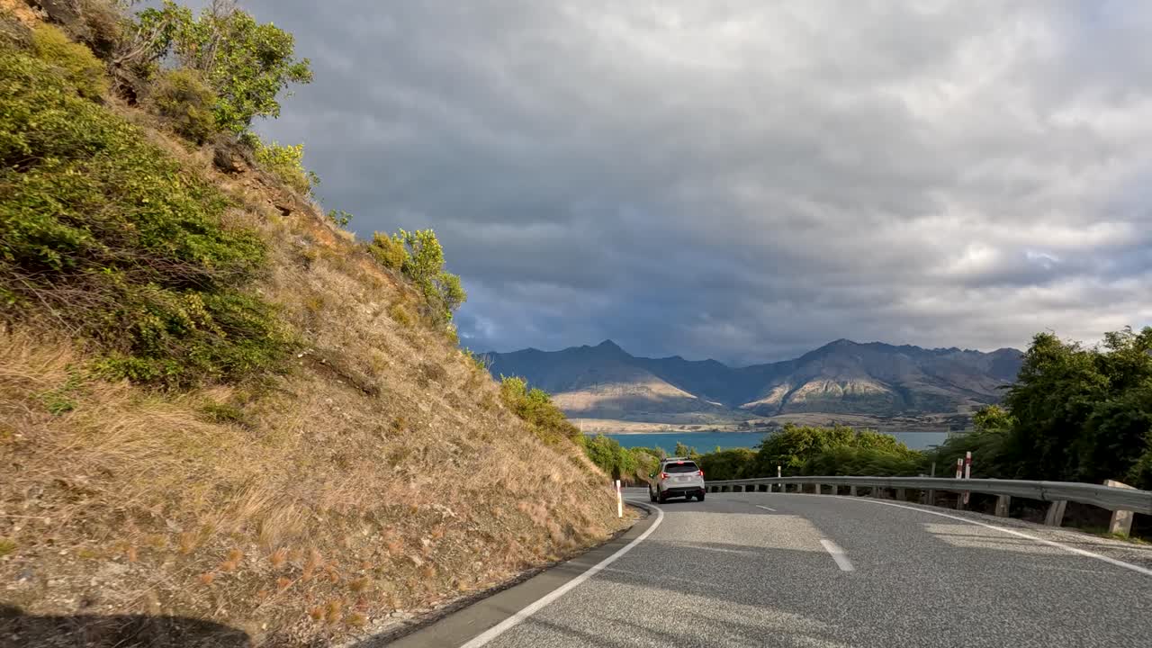 Vehicle travels winding mountain road with lush greenery, dramatic clouds, and distant lake views