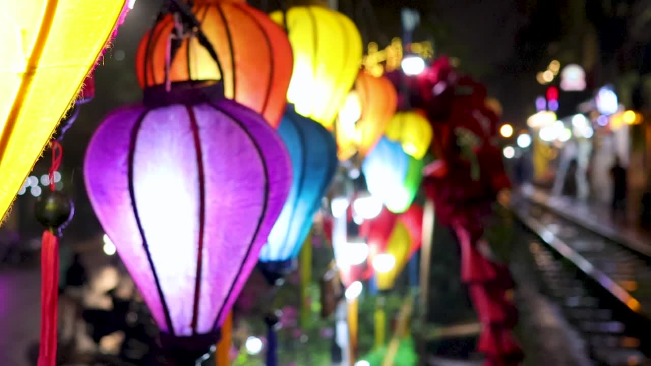 Colorful lanterns illuminate a wet street beside a railway track, creating a vibrant night scene.