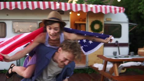 carefree, cheerful couple having fun near trailer in the park, man piggybacking girl with american flag on her back - spinning her around