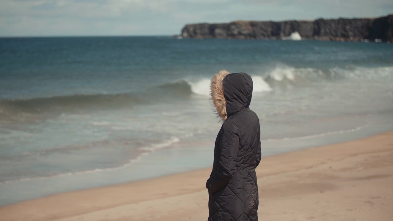 Kvinde i vinterfrakke står på stranden og ser på havets bølger
