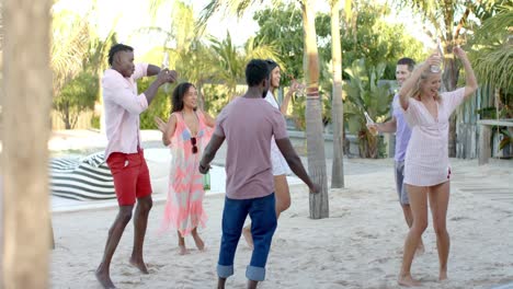 Happy-diverse-group-of-friends-dancing-holding-cocktails-at-beach