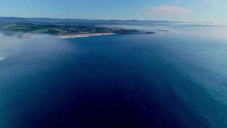 Looking-east-over-the-ocean-from-Kiama-Blowhole-Point,-panning-right-to-left-on-a-glorious-sunny-morning-with-pockets-of-ocean-mist-clinging-to-the-shore-line,-sunlight-reflecting-off-the-calm-sea