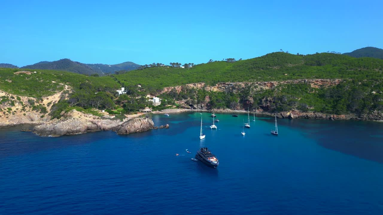 sailboats and yachts moored in turquoise waters of a bay on Ibiza, Spain, surrounded by lush greenery. Fantastic aerial view flight wide orbit overview drone