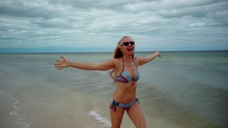 Foto-Frontal-De-Una-Juguetona-Mujer-Rubia-Madura-Con-Gafas-De-Sol-Y-Bikini-Corriendo-Por-La-Orilla-Riéndose-Y-Levantando-Los-Brazos-En-El-Aire-Mostrando-Alegría