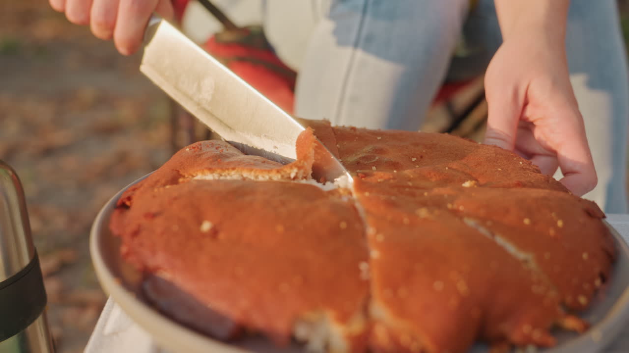 Camping cake being sliced outdoors by camp cook, warm golden light, rustic skillet cake cut into wedges, camp chair and thermos in background, crumbs falling, communal picnic vibe, late afternoon cozy
